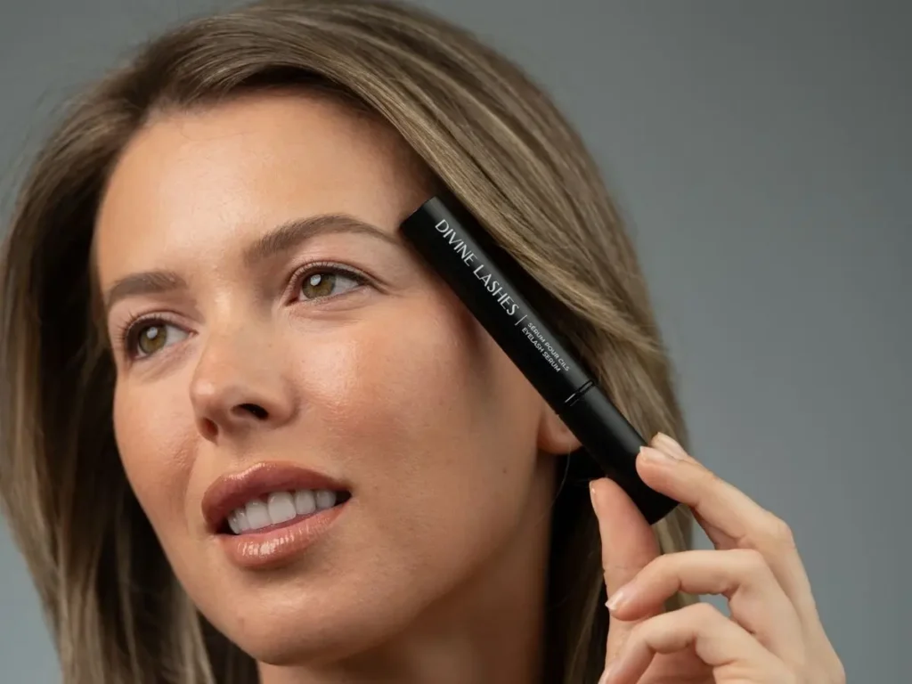 A woman smiling while with the Divine Lashes Eyelash Serum next to her face.
