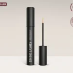 A clean, minimalist studio shot of the black Divine Lashes serum tube and its fine-point applicator brush against a neutral light grey background.