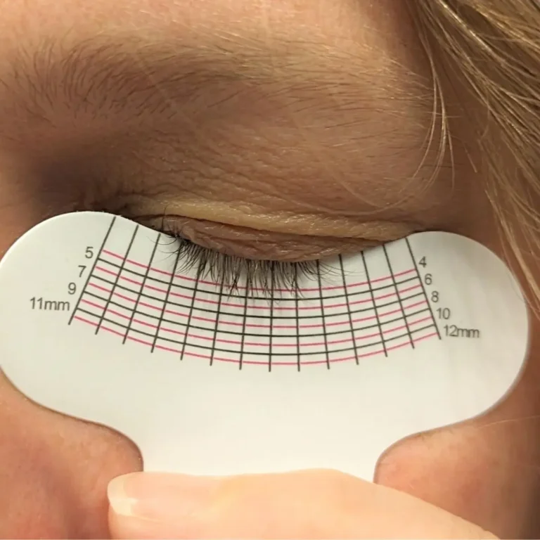 A close-up view of a person's closed eye with light brown hair, showcasing dense and curled eyelashes positioned over a white lash measurement guide from using the Divine Lashes Enhancing Serum.