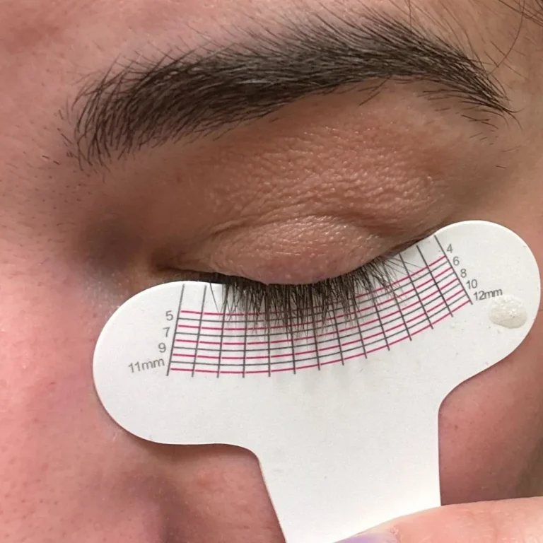 A close-up of a closed eye with dark, thick eyelashes resting against a white semi-circle lash growth measurement tool held against the cheek. Results of the Divine Lashes Eyelash Serum.
